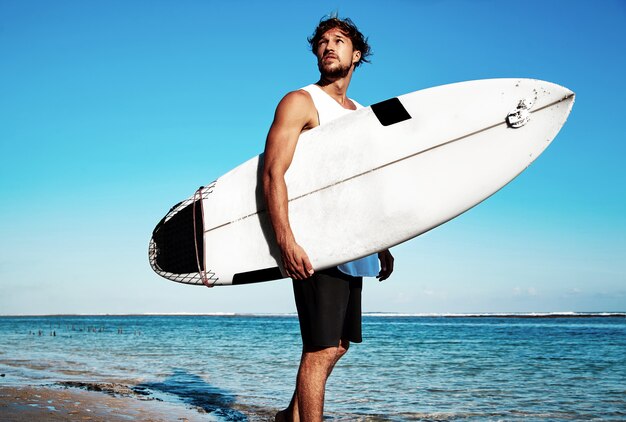 Portret van de knappe hipster zonnebaadde modelsurfer die van de maniermens vrijetijdskleding dragen die met surfplank op blauwe oceaan en hemel gaan