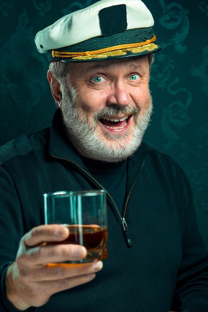 Portret van de oude zeemansmens als kapitein in zwarte sweater en hoed die cognac op zwarte studio drinken