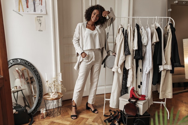 Portret van de volledige lengte van een stijlvolle, donkerhuidige, krullende vrouw in een witte broekjas en een top die zich voordeed in de kleedkamer. Jonge dame leunt op hanger