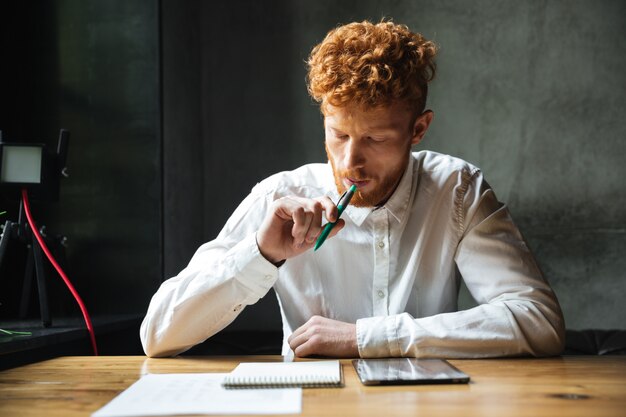 Portret van denken jonge readhead man in wit overhemd, zittend aan houten tafel met pen in zijn mond