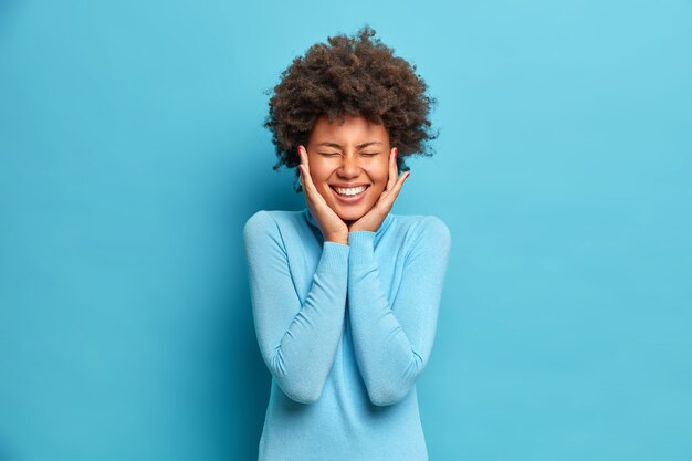 Portret van dolblij Afro-Amerikaanse vrouw met krullend haar houdt de handen op de wangen glimlacht breed