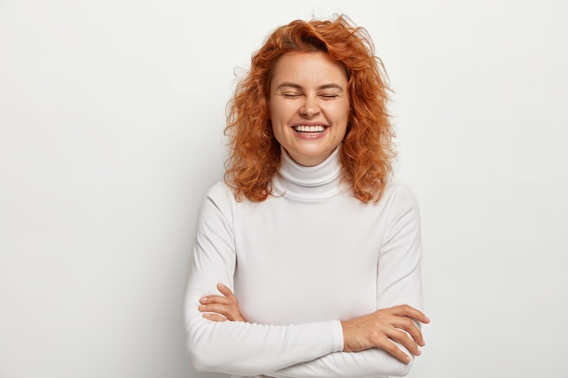 Portret van dolgelukkig gember vrouw lacht uit grappige grap, houdt de armen over elkaar, heeft plezier binnen, houdt de ogen dicht, gekleed in een casual trui, geïsoleerd op wit. Emoties en gevoelens concept.