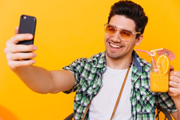 Portret van donkerbruine man die in oranje zonnebril cocktailglas houdt en selfie op oranje ruimte neemt.