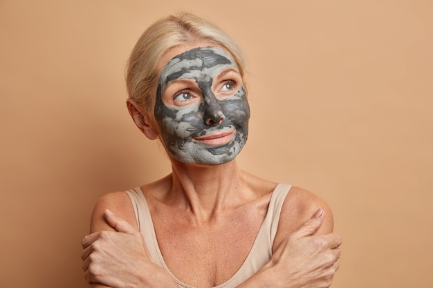 Portret van doordachte senior blanke vrouw kruist armen en raakt schouders zachtjes draagt zelfgemaakte gezichtsmasker thuis geconcentreerd weg geïsoleerd op beige muur