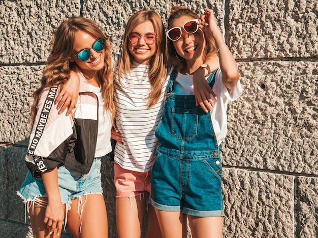 Portret van drie jonge mooie glimlachende hipster meisjes in trendy zomerkleren. Sexy zorgeloze vrouwen poseren in de buurt van muur in de straat. Positieve modellen plezier in zonnebril