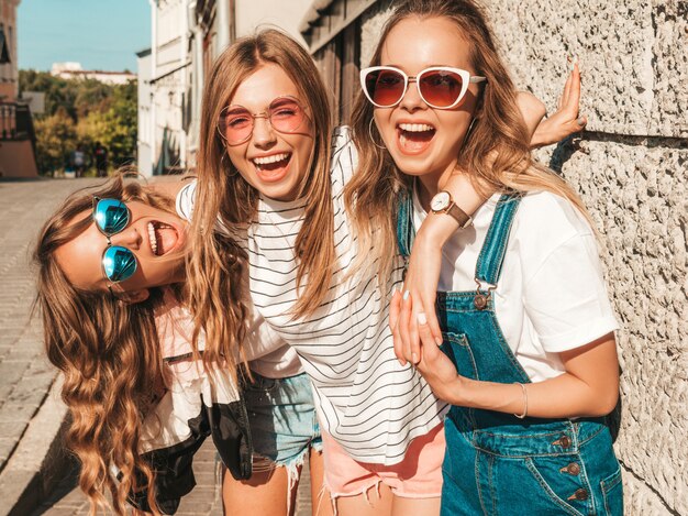 Portret van drie jonge mooie glimlachende hipster meisjes in trendy zomerkleren. Sexy zorgeloze vrouwen poseren in de buurt van muur in de straat. Positieve modellen plezier in zonnebril