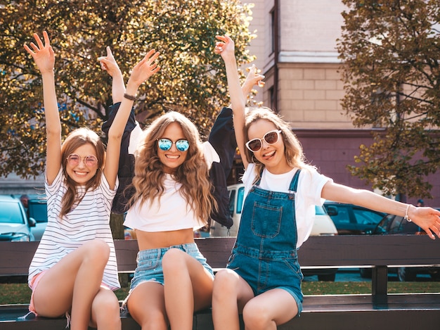 Portret van drie jonge mooie lachende hipster meisjes in trendy zomerkleding. Sexy zorgeloze vrouwen zitten op de bank in de straat. Positieve modellen plezier in zonnebril. Het opheffen van handen