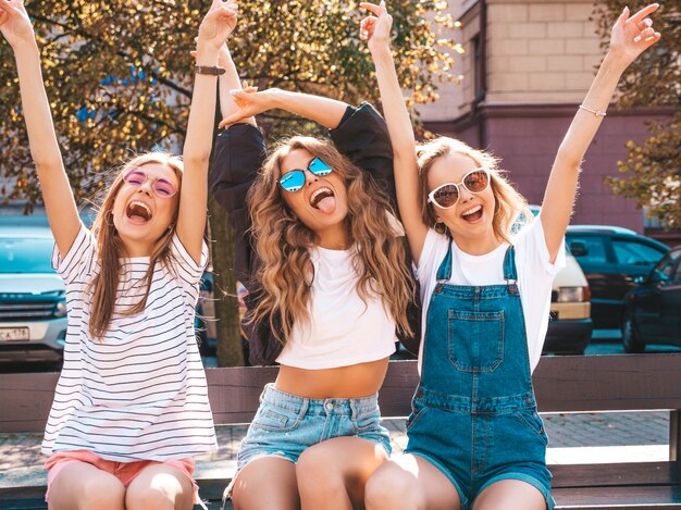 Portret van drie jonge mooie lachende hipster meisjes in trendy zomerkleding. Sexy zorgeloze vrouwen zitten op de bank in de straat. Positieve modellen plezier in zonnebril. Het opheffen van handen