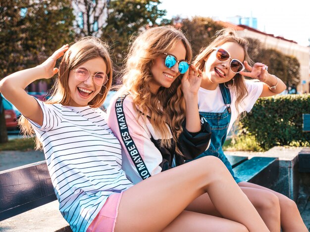 Portret van drie jonge mooie lachende hipster meisjes in trendy zomerkleding. Sexy zorgeloze vrouwen zitten op de bank in de straat. Positieve modellen plezier in zonnebril
