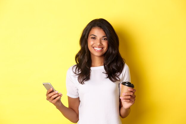 Portret van een aantrekkelijk Afro-Amerikaans meisje dat lacht, koffiekopje en mobiele telefoon vasthoudt, staande op een gele achtergrond.