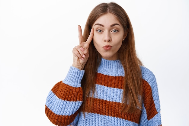 Portret van een aantrekkelijke blanke vrouw die lippen tuit, een kussend gezicht maakt en een v-teken vredesgebaar toont in de buurt van de wang, kawaii poserend, staande over een witte muur