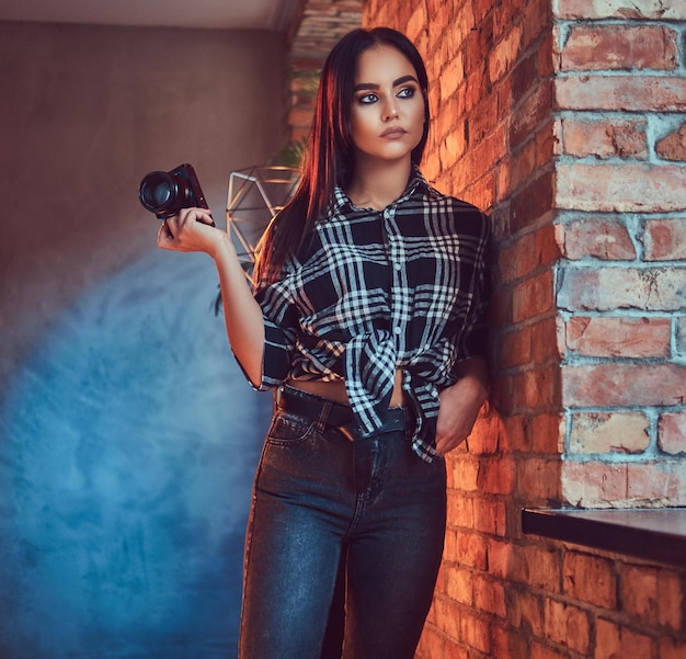 Portret van een aantrekkelijke brunette meisje gekleed in een flanellen shirt en jeans met camera leunend op de muur in een kamer met loft interieur.