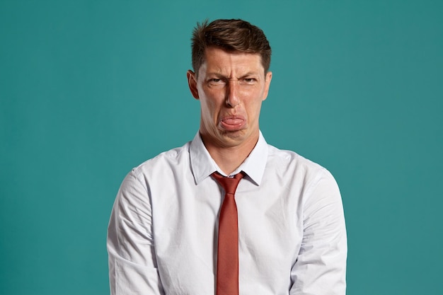 Portret van een aantrekkelijke donkerbruine man met bruine ogen, gekleed in een klassiek wit overhemd en een rode stropdas. Hij doet alsof hij iets niet leuk vindt terwijl hij poseert in een studio tegen een blauwe achtergrond. Conce