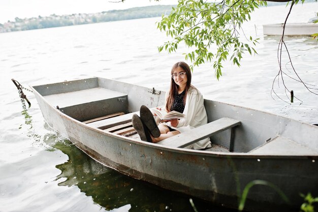 Portret van een aantrekkelijke vrouw met zwarte stippen, een witte sjaal en een bril die een boek leest in een boot op een meer