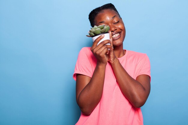 Portret van een afro-amerikaanse jonge vrouw met een bloempot die lacht naar de camera die in de studio staat met een blauwe achtergrond. glimlachende tiener die botanische installatiepot toont. tuinieren concept