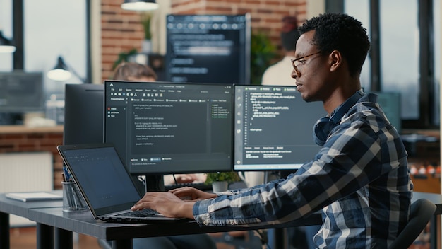 Portret van een afro-amerikaanse ontwikkelaar die een laptop gebruikt om code te schrijven die aan een bureau zit met een algoritme voor het parseren van meerdere schermen in een softwarebureau. coder werkt aan gebruikersinterface met behulp van draagbare computer.