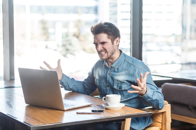 Portret van een agressieve ongelukkige jonge zakenman in een spijkerbroek zit in een café en heeft een slecht humeur en waarschuwt een werknemer via een webcam met opgeheven armen en op elkaar geklemde tanden.
