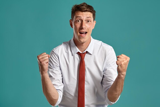 Portret van een atletische donkerbruine man met bruine ogen, gekleed in een klassiek wit overhemd en een rode stropdas. Hij doet alsof hij iets heeft gewonnen in een studio tegen een blauwe achtergrond. Concept van gebaren