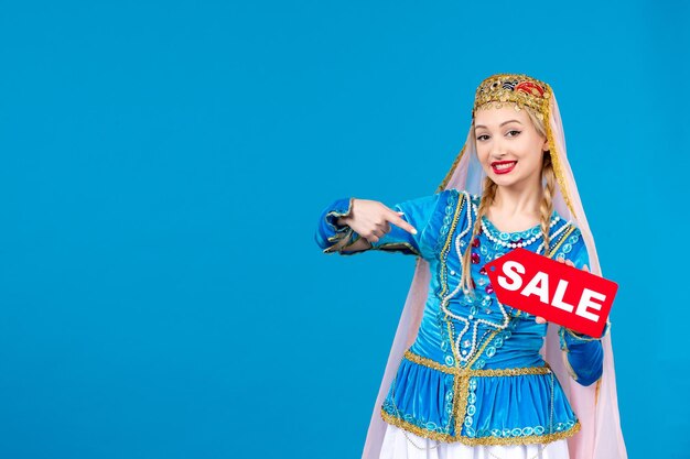 Portret van een azeri-vrouw in traditionele kleding met een rood verkoopnaambordje op een blauwe etnische kleur als achtergrond, novruz shopping lente Gratis Foto