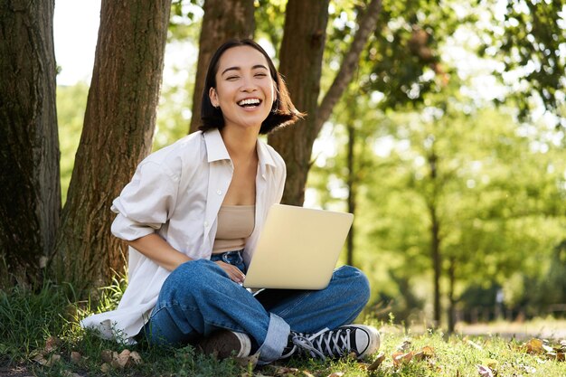 Portret van een Aziatisch meisje met een laptop die op een zonnige zomerdag in de buurt van een boom in het park zit en aan e