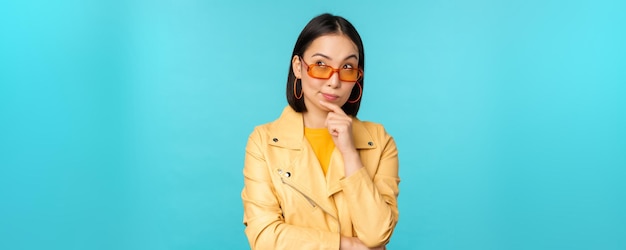 Portret van een aziatische vrouw die denkt op zoek naar doordachte zoekideeën of een oplossing met een zonnebril op een blauwe achtergrond