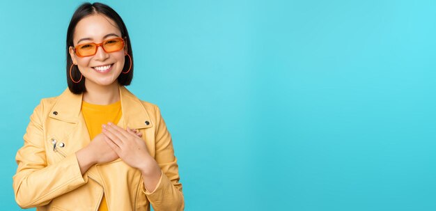 Portret van een aziatische vrouw die lacht, hand in hand op het hart en met tederheid kijkt naar camera dankbare emotie die over blauwe achtergrond staat