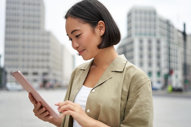 Portret van een aziatische vrouw die leest met een tablet terwijl ze op straat staat te glimlachen terwijl ze naar puin kijkt