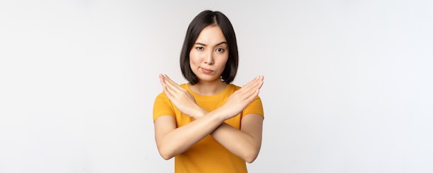 Portret van een aziatische vrouw die serieus en boos kijkt en laat zien dat het taboe-teken van het gebaar verboden is en iets verbiedt dat in gele t-shirt staat op een witte achtergrond