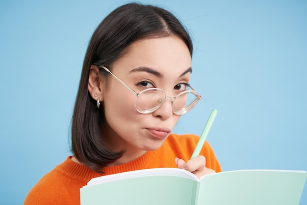 Portret van een aziatische vrouw met een bril die aantekeningen maakt in een notitieboekje, ziet er nieuwsgierig en achterdochtig uit naar camera's
