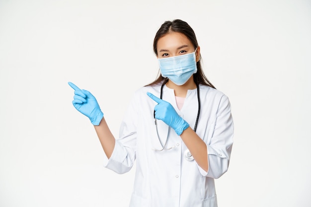 Portret van een aziatische vrouwelijke medische werker die met de vingers naar links wijst, een gezichtsmasker en rubberen handschoenen draagt, die in een kliniekuniform staat op een witte achtergrond