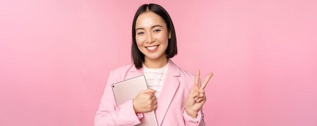 Portret van een Aziatische zakenvrouw in een pak die staat met een digitale tablet die vrede vsign toont en een roze achtergrond glimlacht