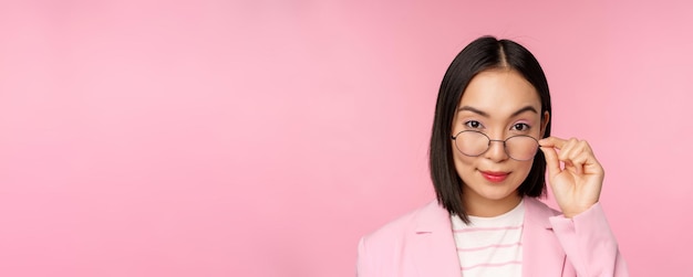 Portret van een aziatische zakenvrouw met een bril die geïntrigeerd naar de camera kijkt en glimlachende professionele verkoopster staart met interesse roze achtergrond