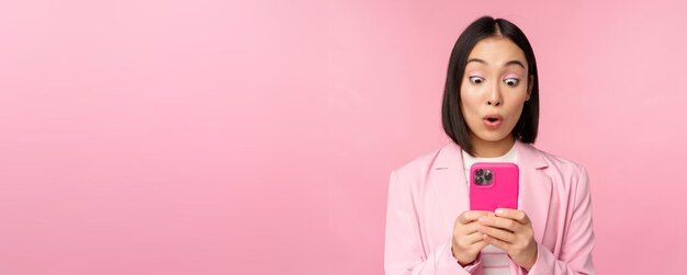 Portret van een Aziatische zakenvrouw met een verbaasd gezicht die een smartphone-app gebruikt in een pak Koreaans meisje met een mobiele telefoon en een opgewonden gezichtsuitdrukking roze achtergrond