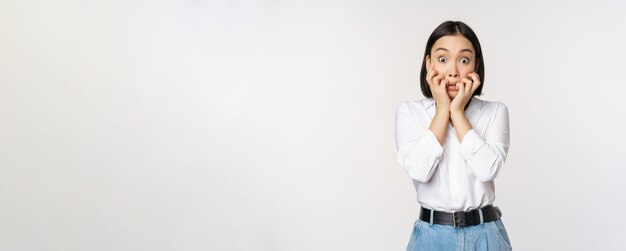 Portret van een bange aziatische vrouw die naar iets eng bijtende vingers op handen kijkt en bang naar de camera kijkt terwijl ze op een witte achtergrond staat