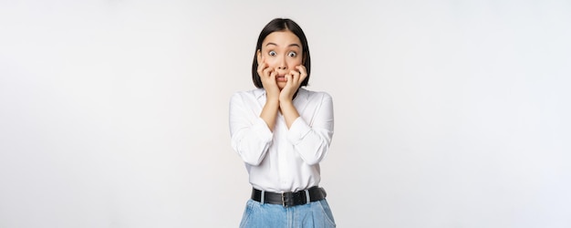 Portret van een bange aziatische vrouw die naar iets eng bijtende vingers op handen kijkt en bang naar de camera kijkt terwijl ze op een witte achtergrond staat