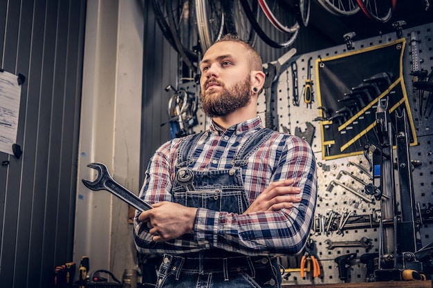 Portret van een bebaarde fietsenmaker met gekruiste armen houdt de bekersleutel vast op de achtergrond van een gereedschapsstandaard in een werkplaats.