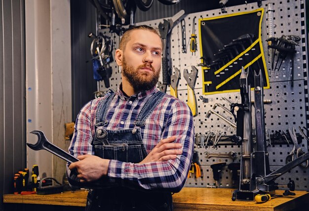 Portret van een bebaarde fietsenmaker met gekruiste armen houdt de bekersleutel vast op de achtergrond van een gereedschapsstandaard in een werkplaats.