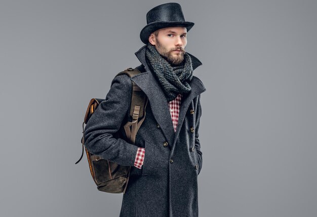 Portret van een bebaarde hipster man gekleed in een grijze jas en een cilinder hoed houdt een rugzak geïsoleerd op een grijze achtergrond.