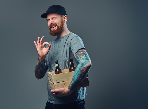 Portret van een bebaarde man in een pet met tatoeages op zijn armen houdt een houten kist met bierflesjes vast.