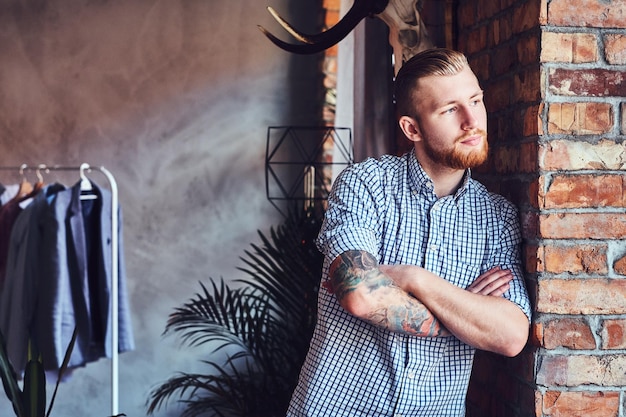 Portret van een bebaarde moderne man met tatoeages op zijn armen poseren bij het raam in een kamer met loft interieur.