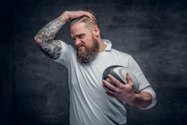 Portret van een bebaarde rugbyspeler met tatoeages op zijn armen houdt een wedstrijdbal vast.