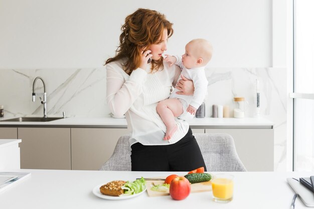 Portret van een bedachtzame moeder die haar schattige kleine baby vasthoudt terwijl ze op haar mobiel praat en in de keuken kookt