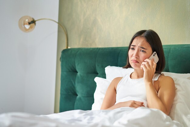 Gratis foto portret van een bezorgde aziatische vrouw die een mobiele telefoon vasthoudt, krijgt een slecht telefoontje en ziet er bezorgd en overstuur uit met een moeilijk telefoongesprek terwijl ze in bed ligt