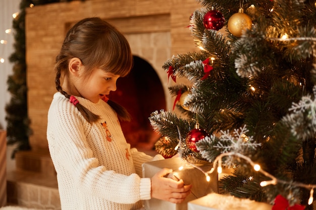 Portret van een blank meisje dat in de buurt van de kerstboom staat en dozen presenteert, een geklede witte trui, donker haar en staartjes, prettige kerstdagen en een gelukkig nieuwjaar.