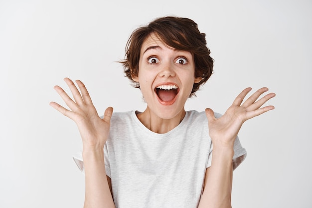 Portret van een blije en verraste vrouw die ja schreeuwt, haar handen verbaasd opsteekt, een supercoole promo-deal bekijkt, staande op een witte muur