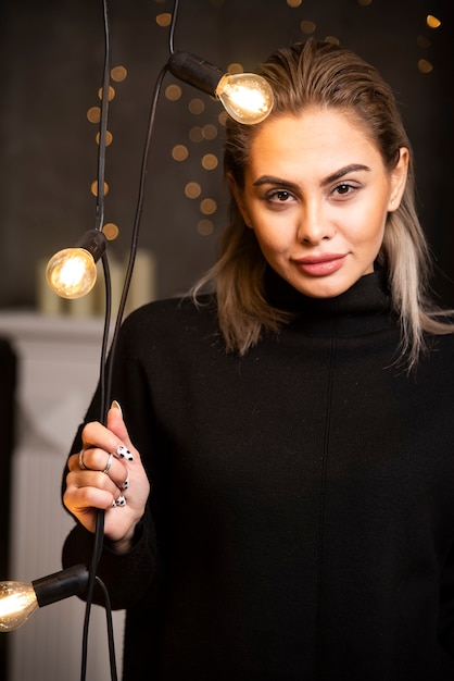 Portret van een blije mooie vrouw in een donkere trui die in de buurt van lampen staat.