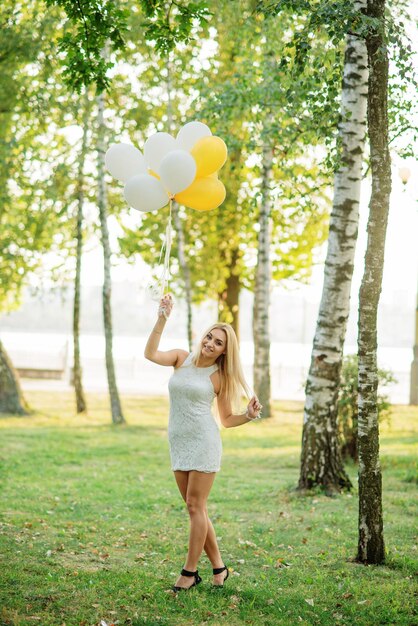 Portret van een blond meisje in een witte jurk met ballonnen bij handen tegen het park op een vrijgezellenfeestje