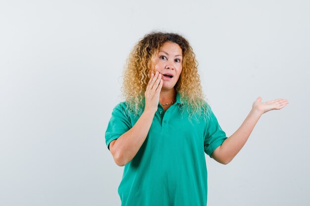 Portret van een blonde dame met krullend haar die de hand op de wang houdt, de handpalm opzij spreidt in een groen T-shirt en er verbaasd uitziet aan de voorkant