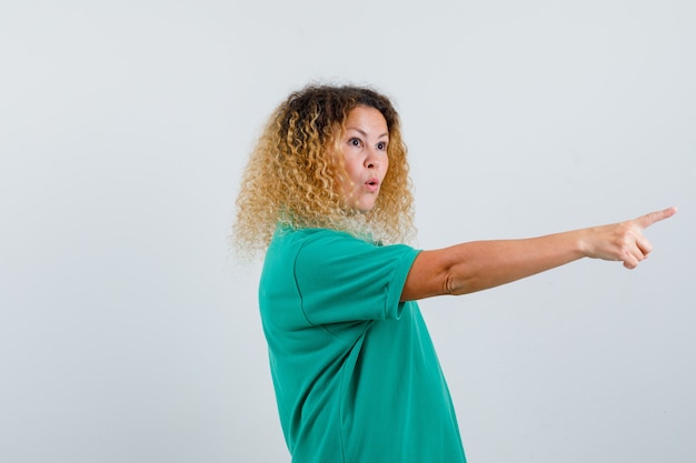 Portret van een blonde dame met krullend haar die wegwijst in een groen t-shirt en een verbaasd vooraanzicht kijkt