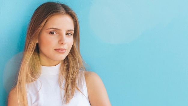 Portret van een blonde jonge vrouw die camera tegen blauwe achtergrond bekijkt